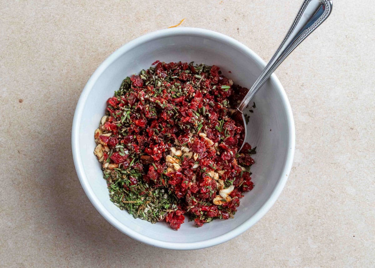 A white mixing bowl filled with chopped dried cranberries, toasted pecans, rosemary, and thyme, stirred together with a silver spoon to make the coating for a goat cheese log.