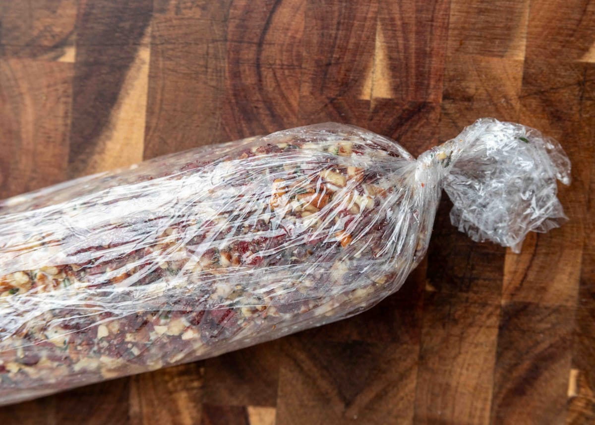 A cranberry pecan goat cheese log wrapped tightly in plastic wrap, resting on a wooden cutting board with visible grain patterns.