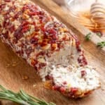 A cranberry pecan goat cheese log on a wooden serving board with one end sliced to reveal the creamy interior. The surface is coated with finely chopped dried cranberries, toasted pecans, and herbs, and the log is drizzled with honey. A honey dipper and fresh rosemary sprigs are visible in the background.