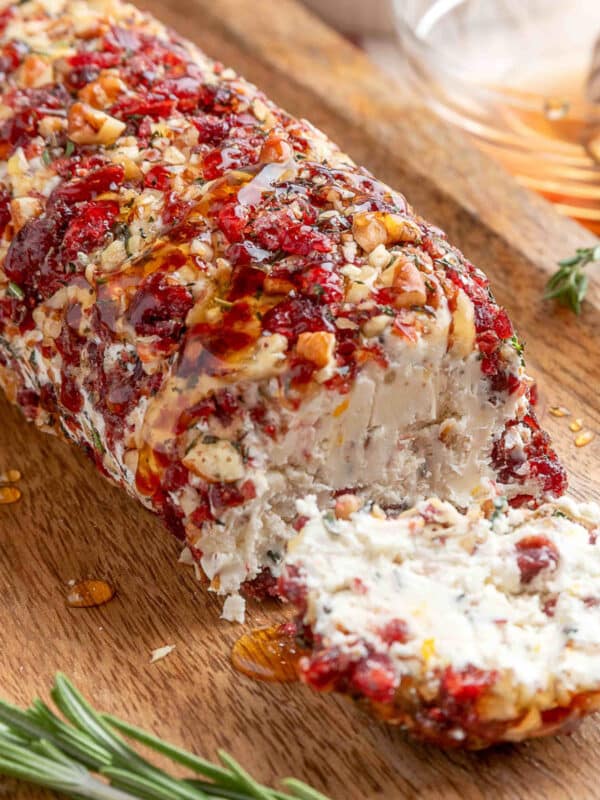 A cranberry pecan goat cheese log on a wooden serving board with one end sliced to reveal the creamy interior. The surface is coated with finely chopped dried cranberries, toasted pecans, and herbs, and the log is drizzled with honey. A honey dipper and fresh rosemary sprigs are visible in the background.