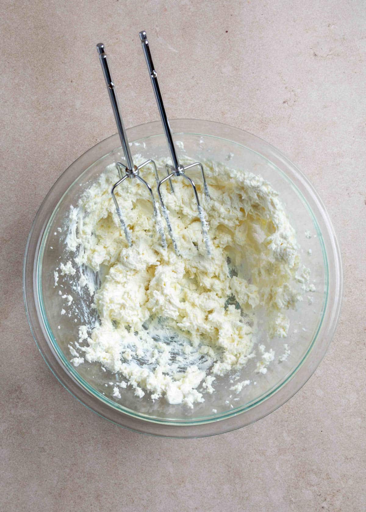 A glass mixing bowl filled with whipped goat cheese and cream cheese, with electric mixer beaters resting inside. The mixture appears light and fluffy against a neutral countertop background.