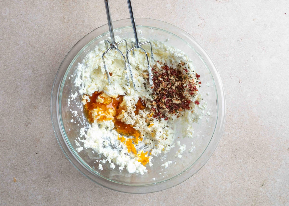 Overhead view of a glass mixing bowl containing softened goat cheese and cream cheese, partially blended with a hand mixer. Honey, black pepper, and a portion of the chopped cranberry-pecan-herb mixture have been added to the bowl.