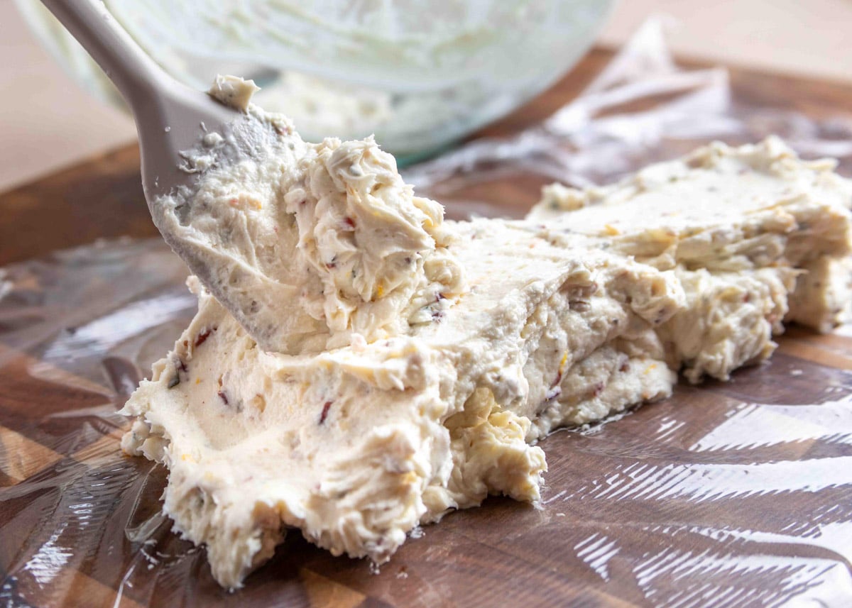 A creamy goat cheese mixture with finely chopped pecans and cranberries is being spooned onto plastic wrap in preparation for forming into a log, with a mixing bowl visible in the background.