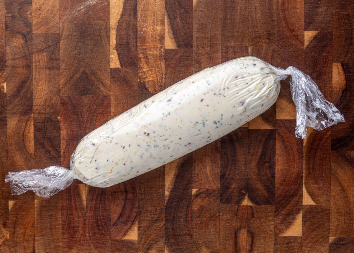 A log-shaped mixture of goat cheese, dried cranberries, pecans, herbs, and orange zest wrapped tightly in plastic wrap on a dark wooden cutting board, ready to be chilled.