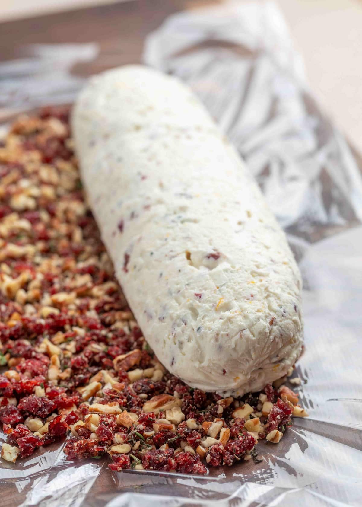 A log of goat cheese mixed with cranberries, orange zest, and herbs being rolled in a chopped coating mixture of toasted pecans, dried cranberries, rosemary, and thyme on plastic wrap.