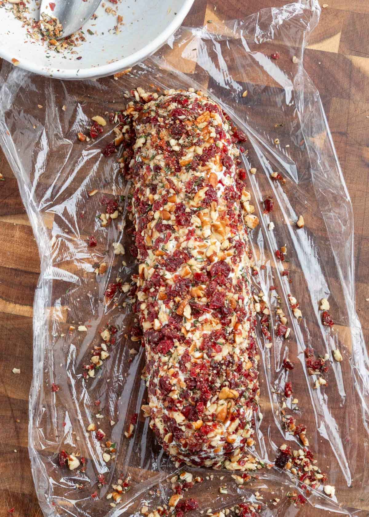 A log of goat cheese mixture coated in finely chopped dried cranberries, toasted pecans, and fresh herbs, resting on a sheet of plastic wrap on a wooden cutting board. A white mixing bowl with leftover coating is visible in the corner.