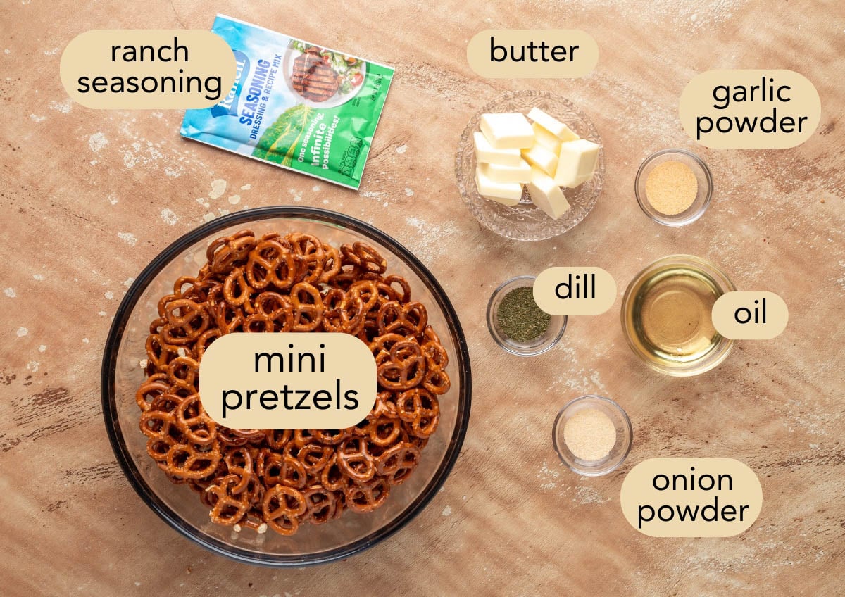 Flat lay of labeled ingredients for ranch pretzels on a tan surface, including a large bowl of mini pretzels, ranch seasoning packet, cubes of butter, garlic powder, onion powder, dried dill, and oil in small glass bowls.