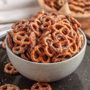 A gray bowl filled with Ranch Pretzels on a black plate, with a second bowl in the background and a few seasoned pretzels scattered on the plate.