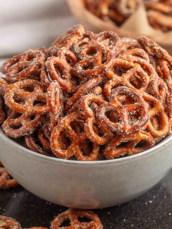 A gray bowl filled with Ranch Pretzels on a black plate, with a second bowl in the background and a few seasoned pretzels scattered on the plate.
