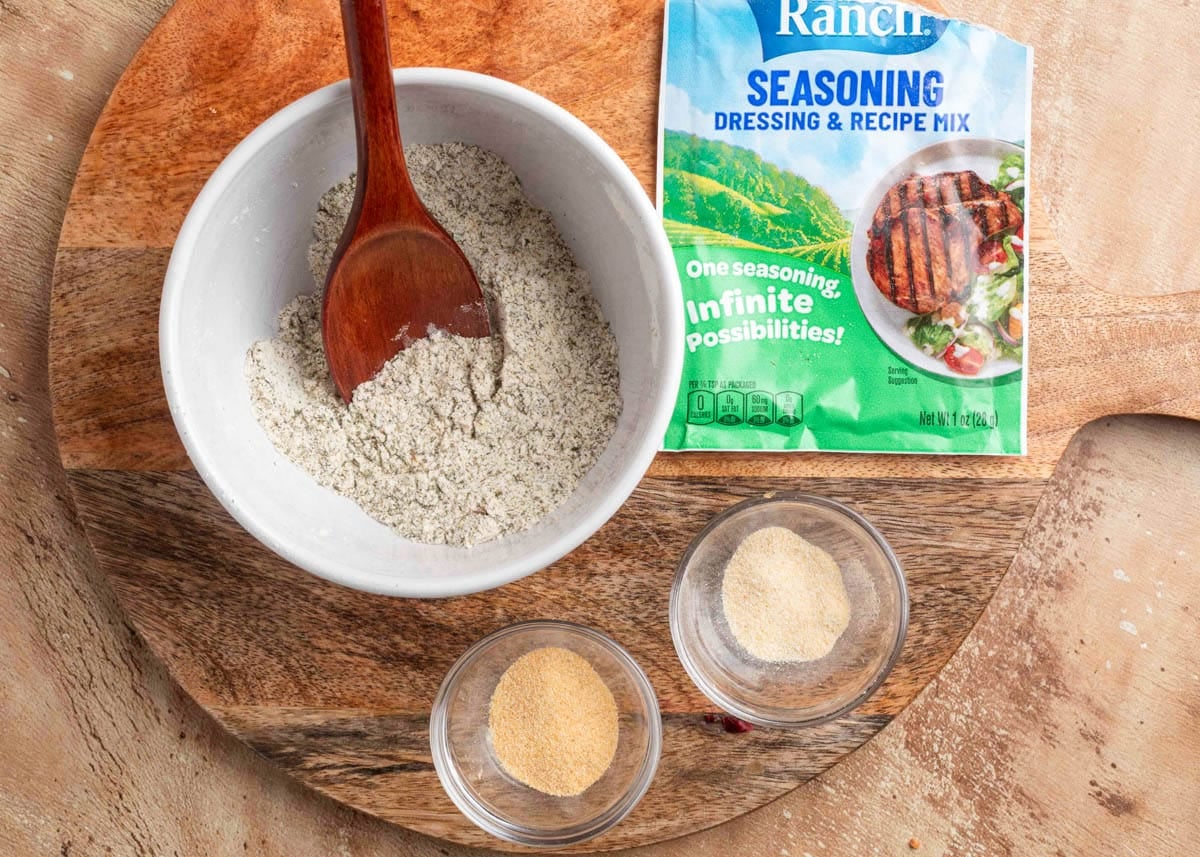 Overhead view of a bowl of ranch seasoning mixture being stirred with a wooden spoon on a round wooden cutting board, surrounded by a ranch dressing mix packet, garlic powder, and onion powder in small glass bowls.