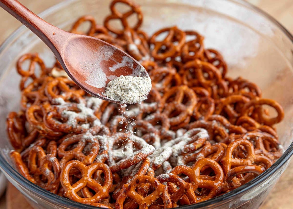 A wooden spoon sprinkles ranch seasoning mix over a large glass bowl filled with mini pretzels coated in melted butter.