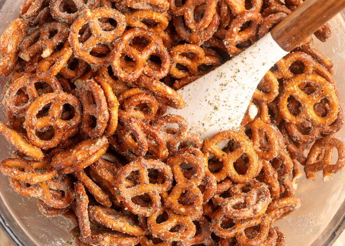 Mini pretzels coated in ranch seasoning are tossed in a glass mixing bowl with a white spatula.