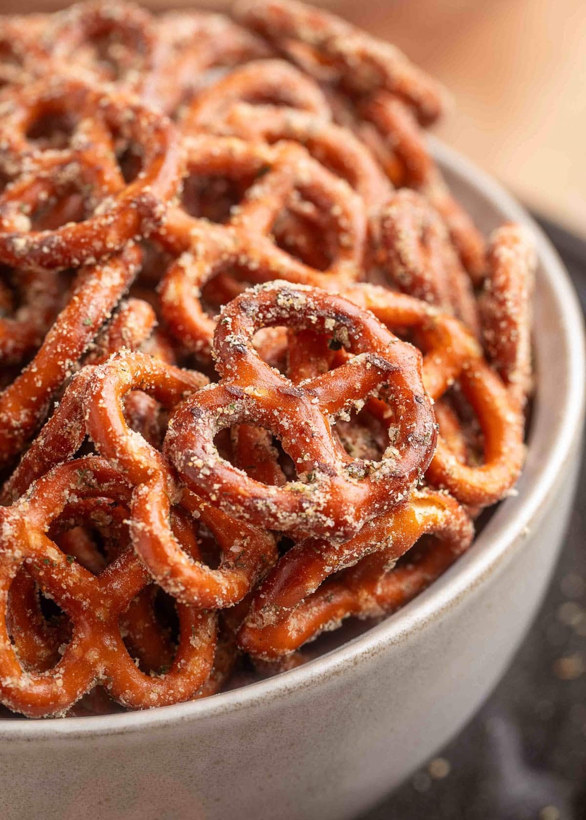 Closeup of seasoned Ranch Pretzels piled high in a beige ceramic bowl, highlighting their crisp texture and coating of dried herbs and spices.