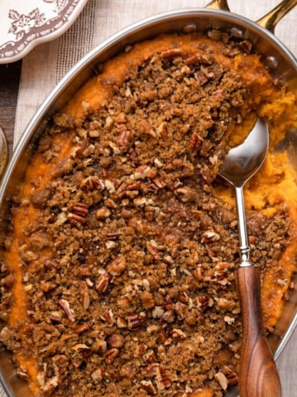 Overhead view of a sweet potato casserole in a stainless steel baking dish with pecan and brown sugar streusel topping and a serving spoon resting in a scooped-out section.
