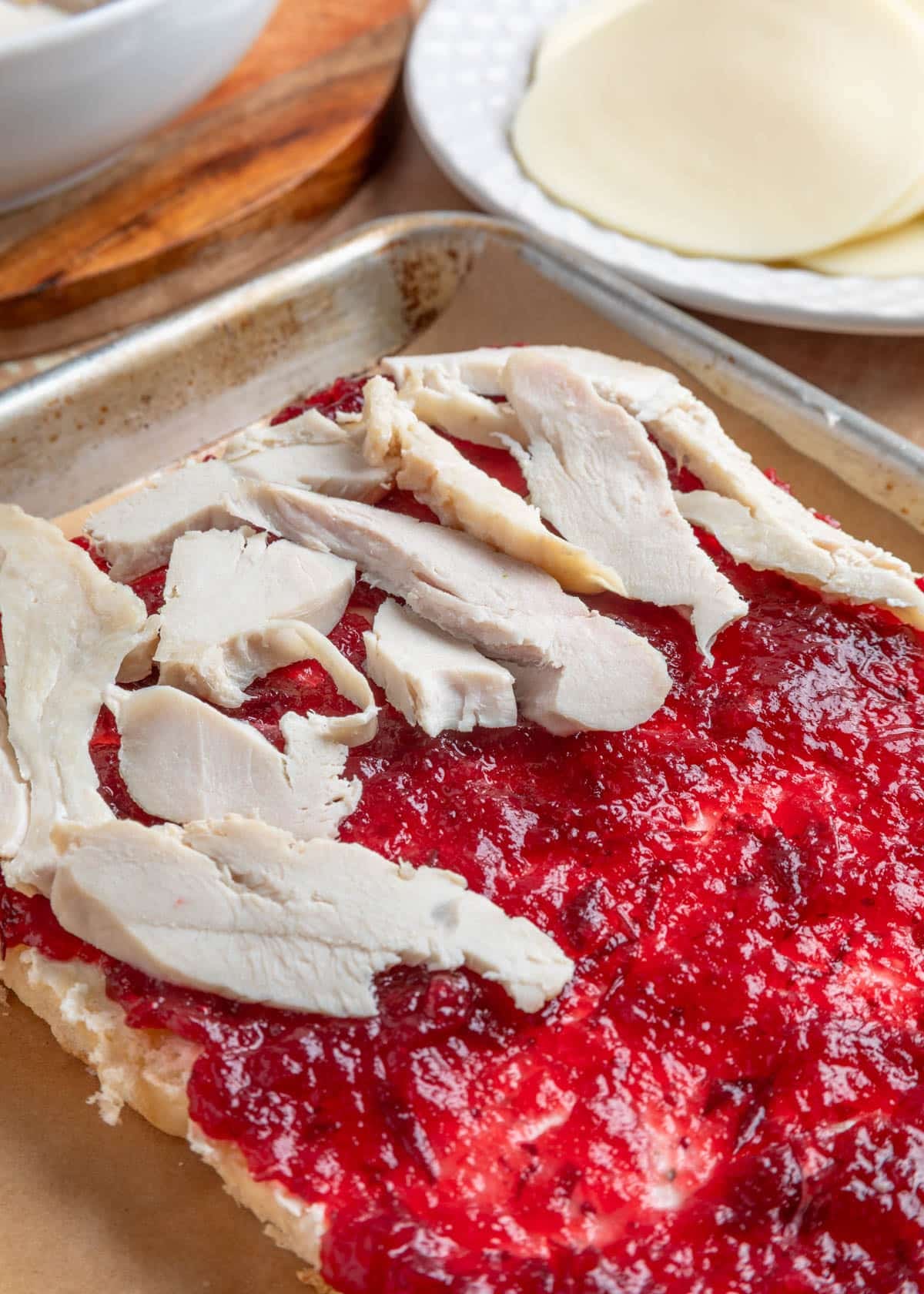 Close-up of sliced turkey layered over cranberry sauce on the bottom half of Hawaiian rolls, set on a parchment-lined sheet pan. Provolone cheese slices and more turkey are visible in the background.
