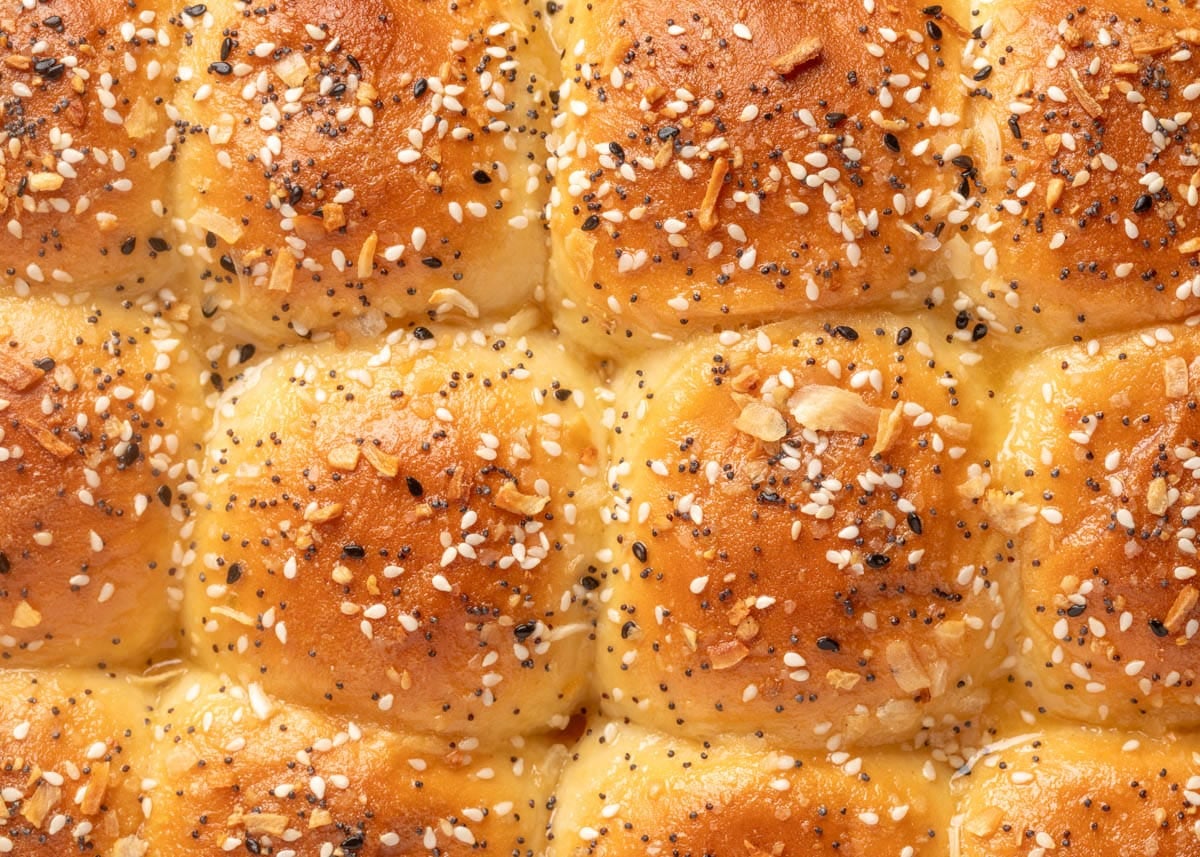 Close-up of golden baked slider buns topped with everything bagel seasoning, including sesame seeds, poppy seeds, dried onion, and garlic flakes.