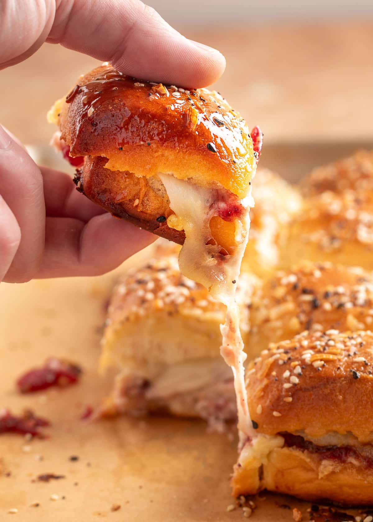 A close-up of a hand lifting a turkey cranberry slider from a baking sheet, with melted provolone cheese stretching from the sandwich. The slider is topped with a golden everything-seasoned roll and baked until glossy.
