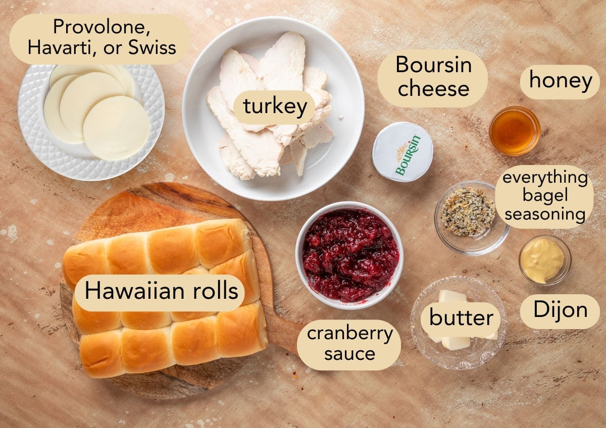 Labeled ingredients for turkey cranberry sliders arranged on a wood-toned surface, including Hawaiian rolls, turkey, provolone cheese, cranberry sauce, Boursin, Dijon, honey, butter, and everything bagel seasoning.