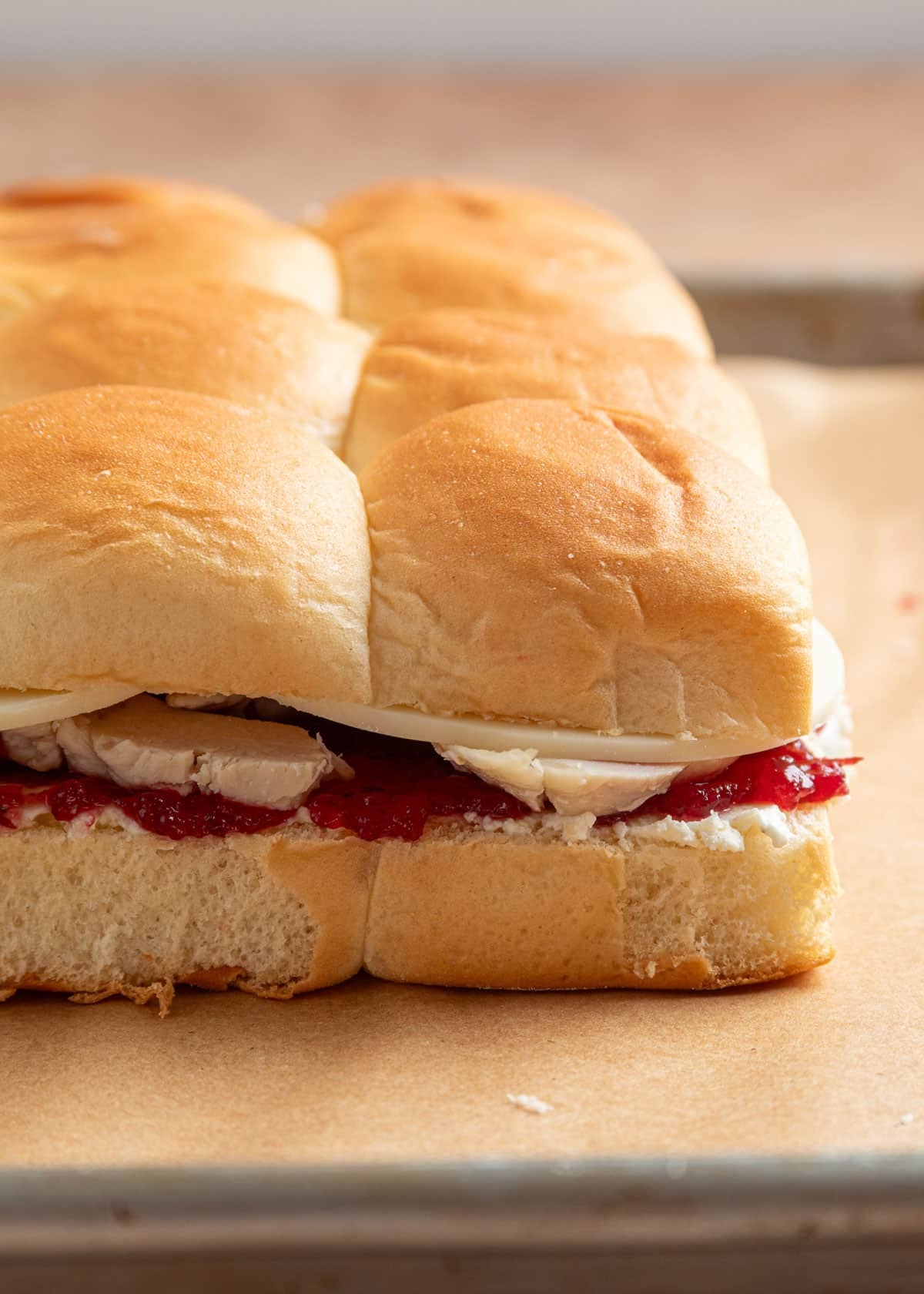 Close-up of unbaked turkey cranberry sliders on a baking sheet, with layers of turkey, cranberry sauce, Boursin, and cheese between halved Hawaiian rolls.