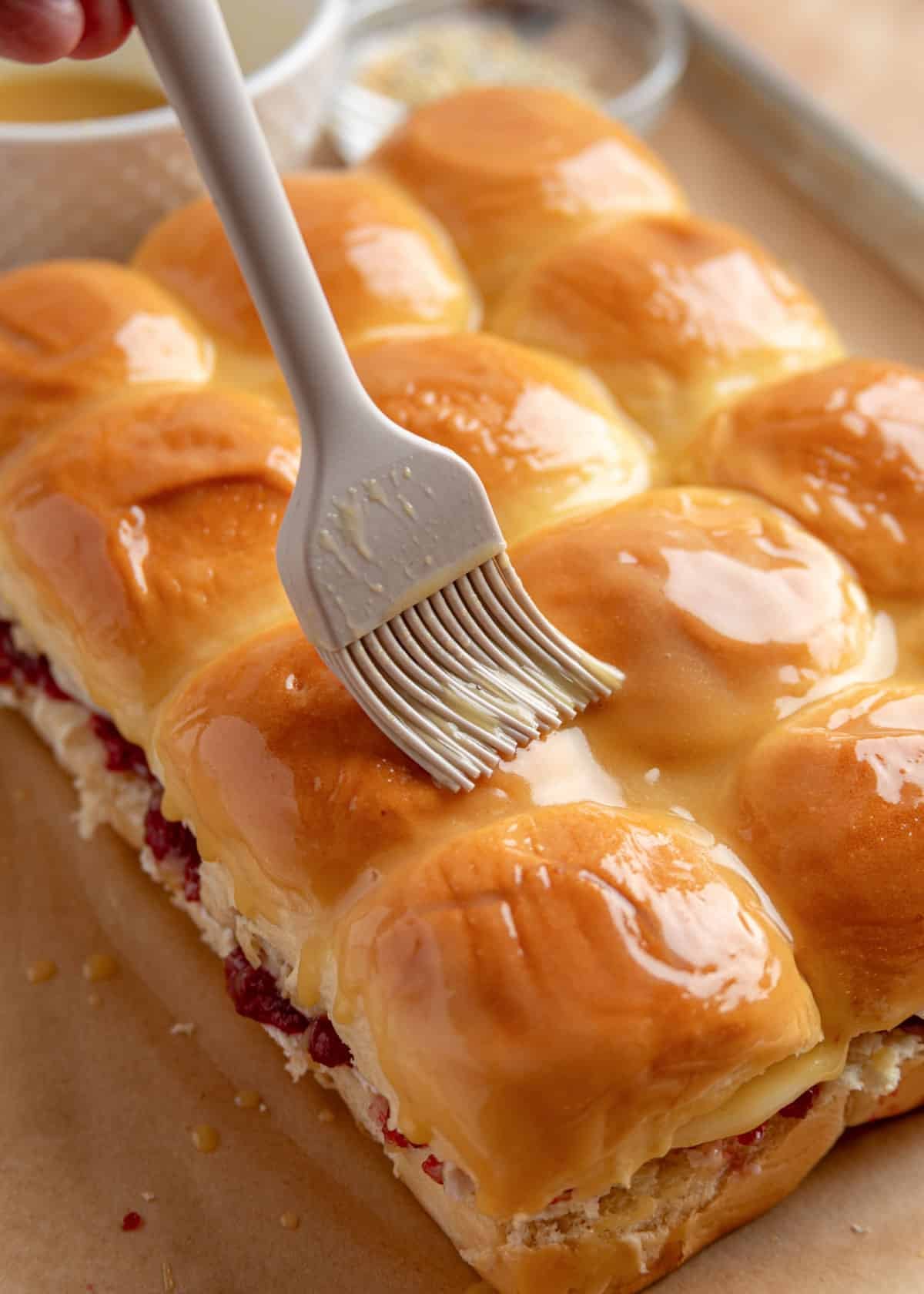 Hawaiian roll slider tops being brushed with a buttery Dijon-honey mixture before baking, with visible cranberry and cheese filling along the edges.
