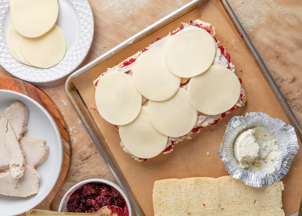 Overhead view of assembled turkey cranberry sliders in progress on a parchment-lined baking sheet, with provolone slices layered over turkey, cranberry sauce, and Boursin. Surrounding ingredients include a dish of sliced turkey, a bowl of cranberry sauce, provolone on a plate, and a foil wrapper of cheese.