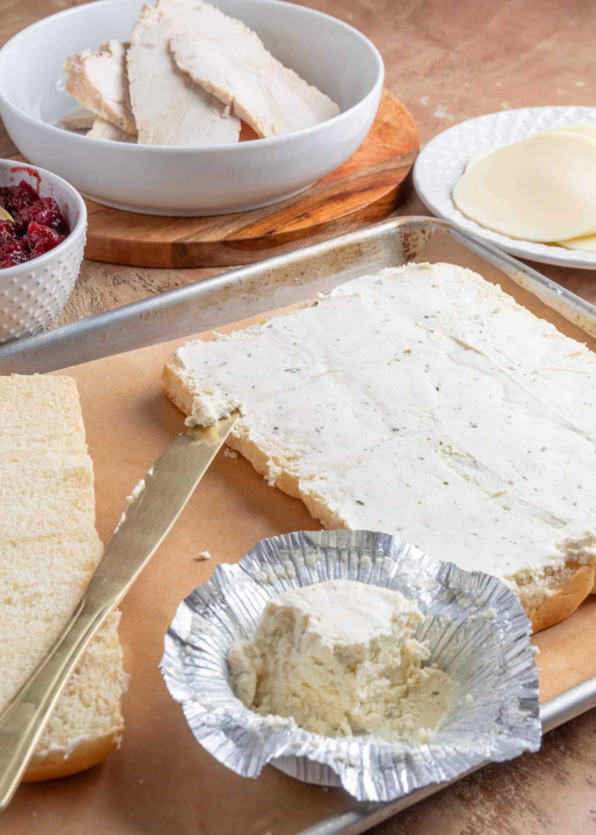 Step-by-step image showing the bottom half of Hawaiian rolls spread with Boursin cheese on a parchment-lined sheet pan. A foil cheese wrapper and spreading knife rest nearby, with bowls of cranberry sauce, turkey slices, and provolone in the background.