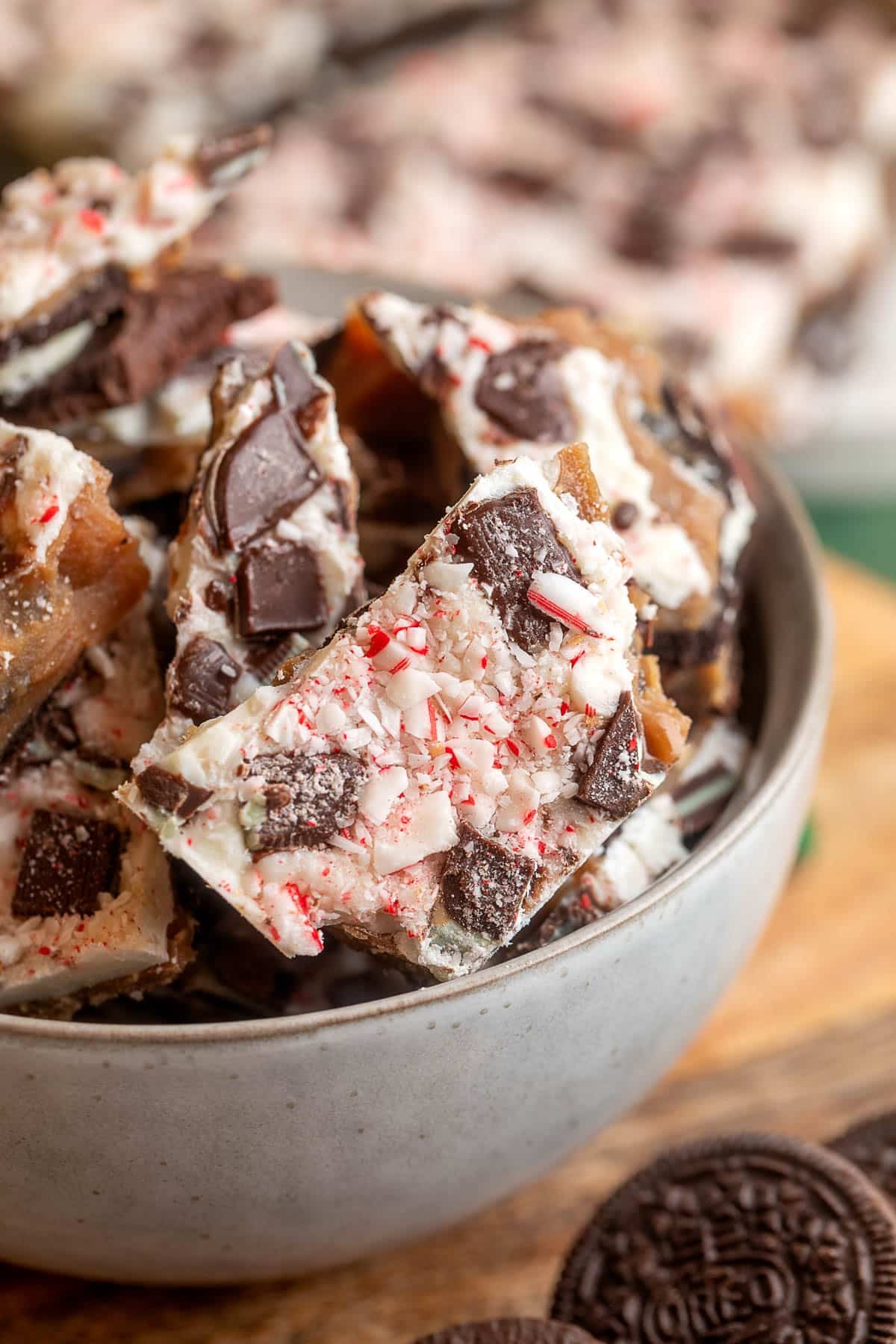 Close-up of Oreo chocolate mint toffee bark in a gray bowl, topped with crushed peppermint candies and chunks of Oreo cookies, with more bark pieces and whole Oreos visible in the background.