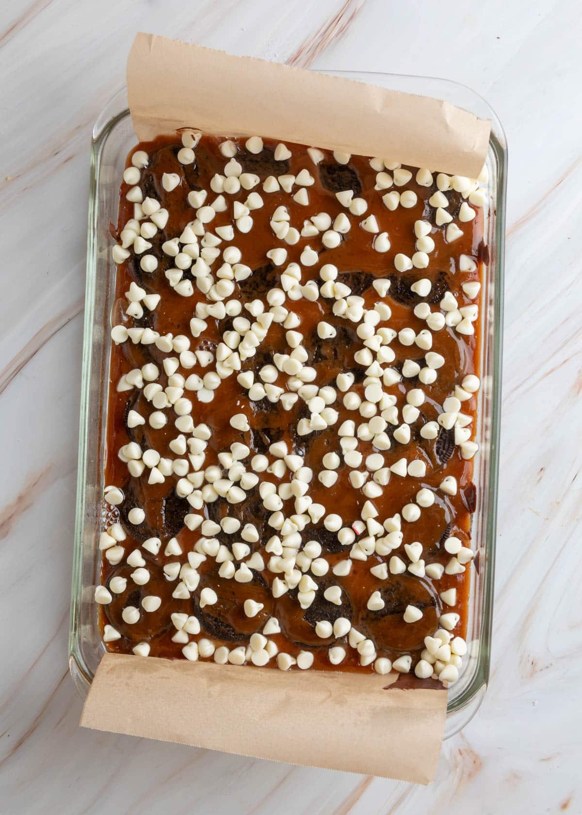 Overhead view of a glass baking dish filled with toffee-covered Oreo halves, topped with scattered white chocolate chips in a parchment-lined pan.