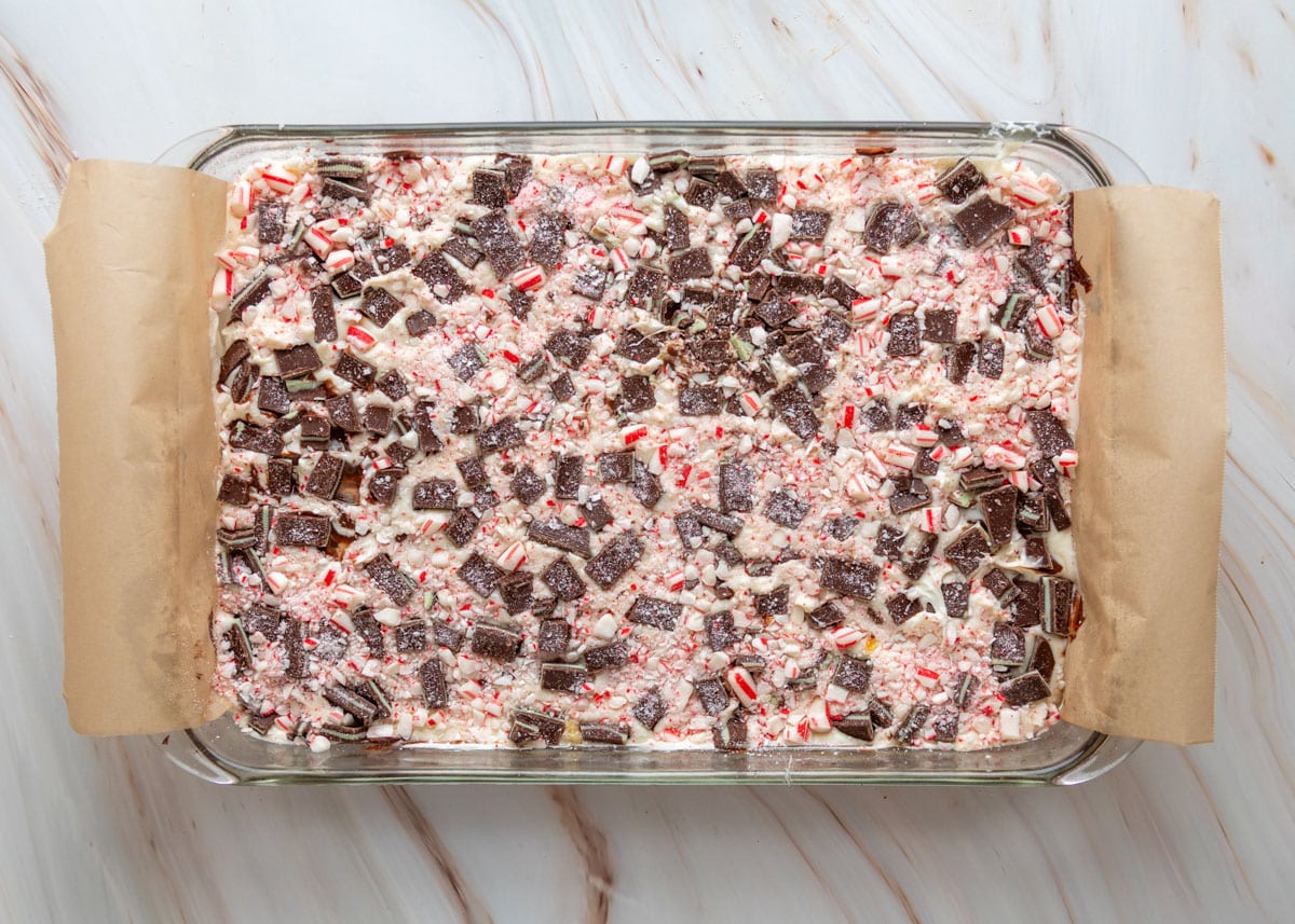 Overhead view of a glass baking dish filled with layered Oreo mint toffee bark, topped generously with chopped Andes mints and crushed peppermint candies on a creamy white chocolate base.