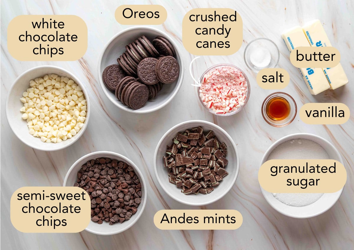 Flat lay of labeled ingredients in bowls for Oreo chocolate mint toffee bark, including white and semisweet chocolate chips, Oreos, crushed candy canes, chopped Andes mints, sugar, butter, vanilla, and salt on a marble surface.