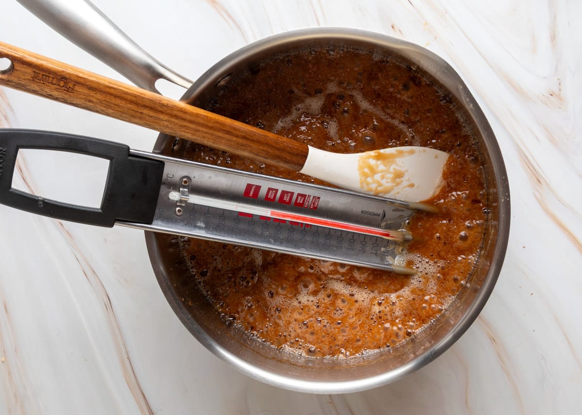 A stainless steel saucepan filled with dark golden toffee is shown bubbling on the stove, with a candy thermometer clipped to the side and a white spatula resting across the top.