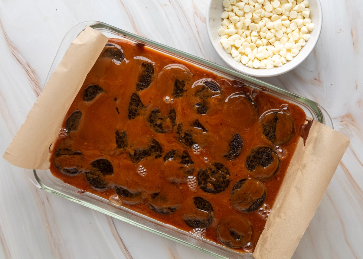 Glass baking dish filled with glossy, bubbling toffee poured over a layer of whole Oreos on top of melted chocolate, with a bowl of white chocolate chips placed nearby.