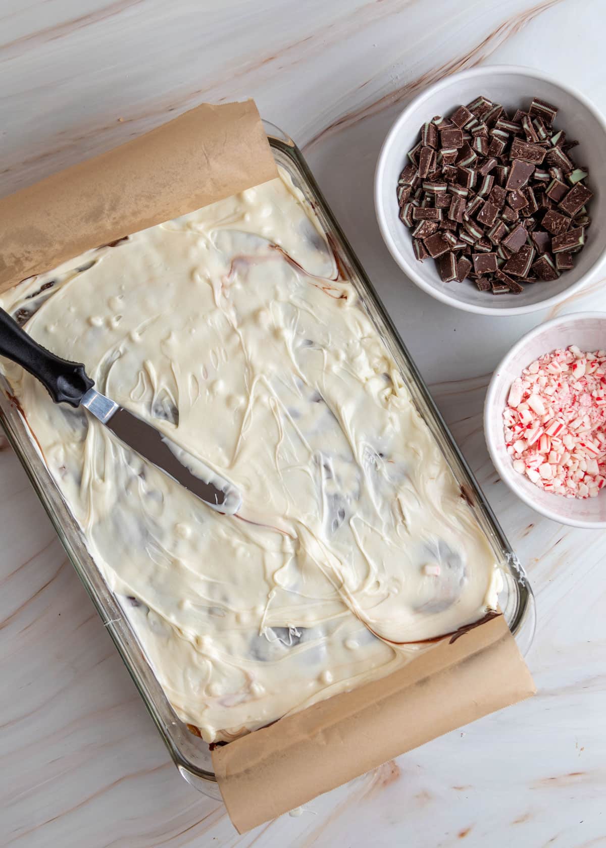 White chocolate spread evenly over the toffee bark in a glass dish with an offset spatula resting on top; bowls of chopped Andes mints and crushed peppermint candies are nearby on the marble surface.