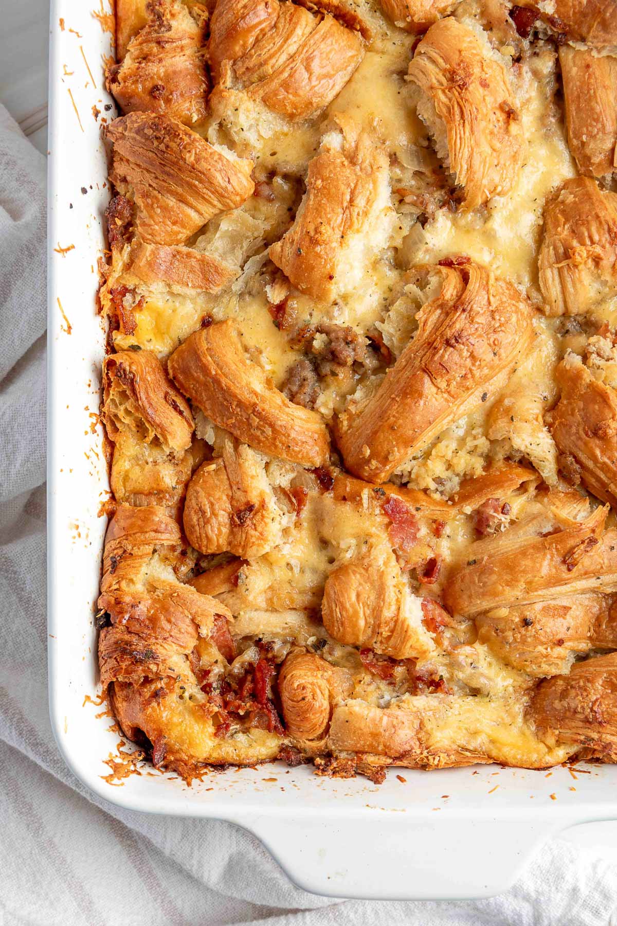 Golden brown croissant pieces baked into a savory sausage breakfast casserole, nestled in a creamy, cheesy egg custard with visible bits of browned sausage, bacon, and crisped edges along the baking dish.