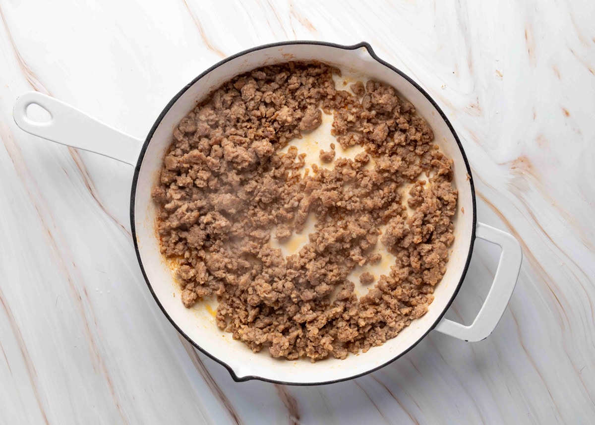 Overhead view of a white enamel skillet filled with crumbled, browned breakfast sausage, freshly cooked and still steaming, on a light marble surface.