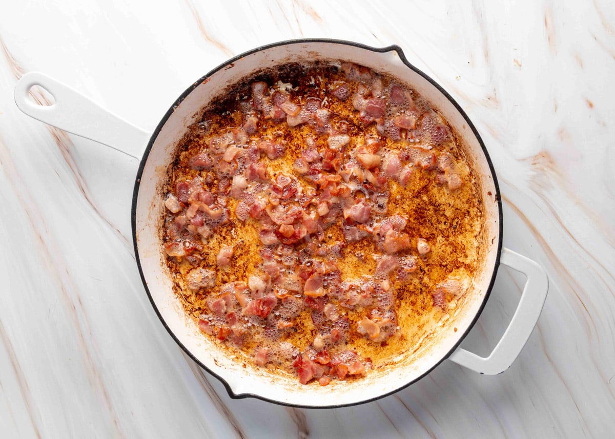 Close-up of chopped bacon sizzling and bubbling in rendered fat in a white enameled skillet with a golden brown fond on the bottom.