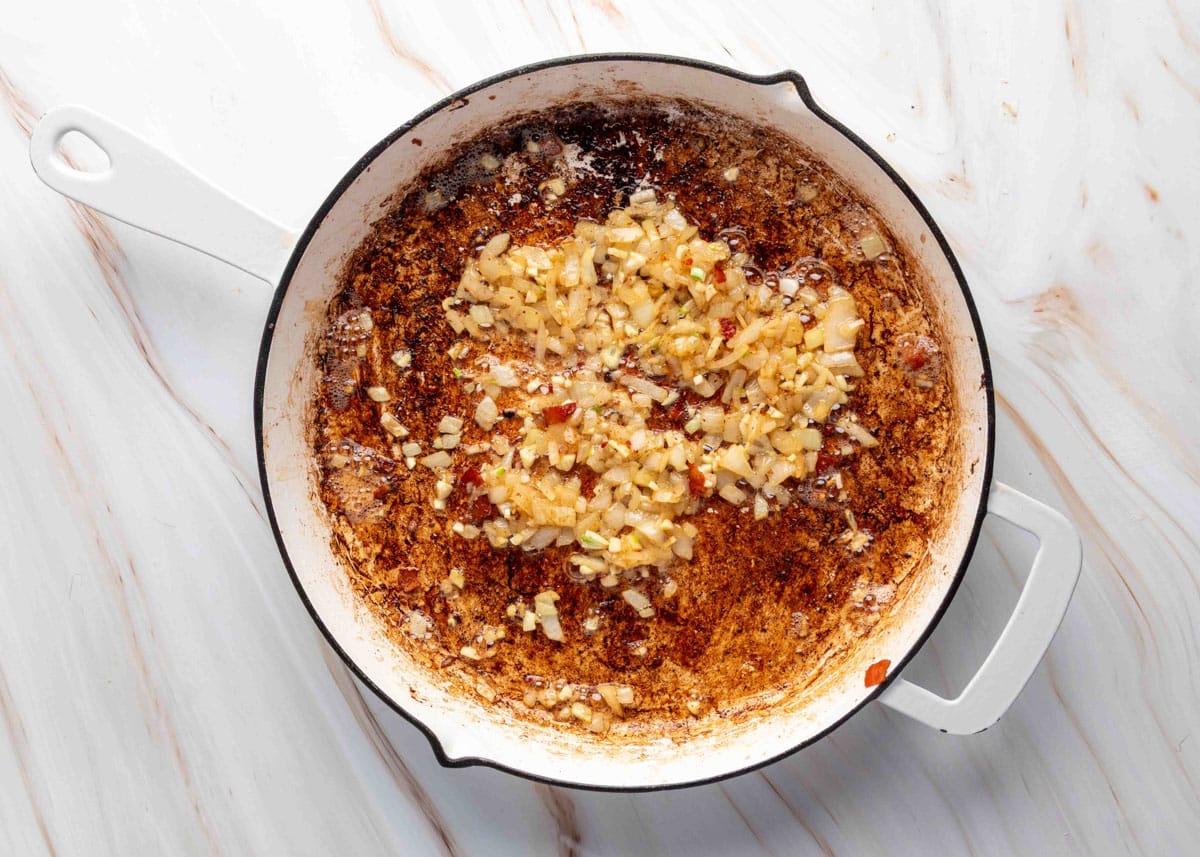 Diced onion and minced garlic cooking in bacon drippings in a white skillet, surrounded by browned bits for added flavor.