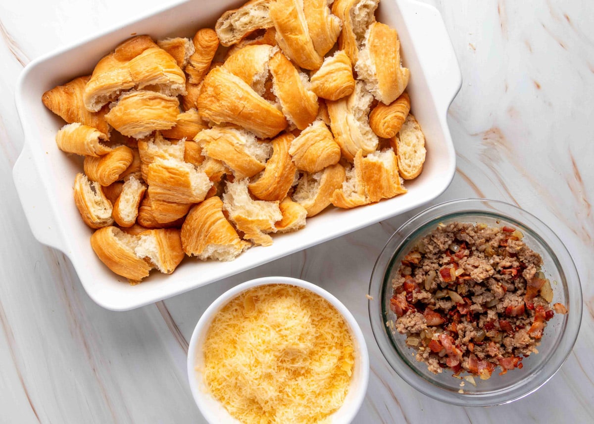 Overhead view of a baking dish filled with torn croissants next to two bowls&mdash;one with shredded cheese, and the other with a cooked mixture of sausage, bacon, and onions&mdash;showing the components for assembling a breakfast casserole.