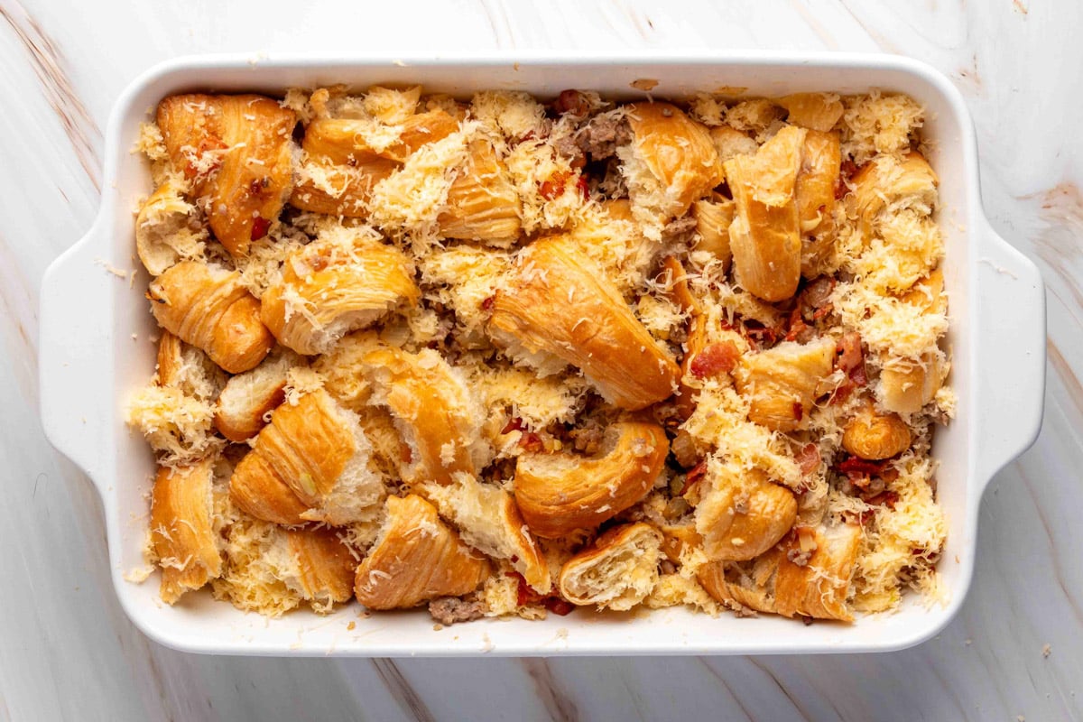 A white rectangular baking dish filled with torn croissants, cooked sausage, crispy bacon, and shredded cheese, layered together before adding the egg mixture for a breakfast casserole.