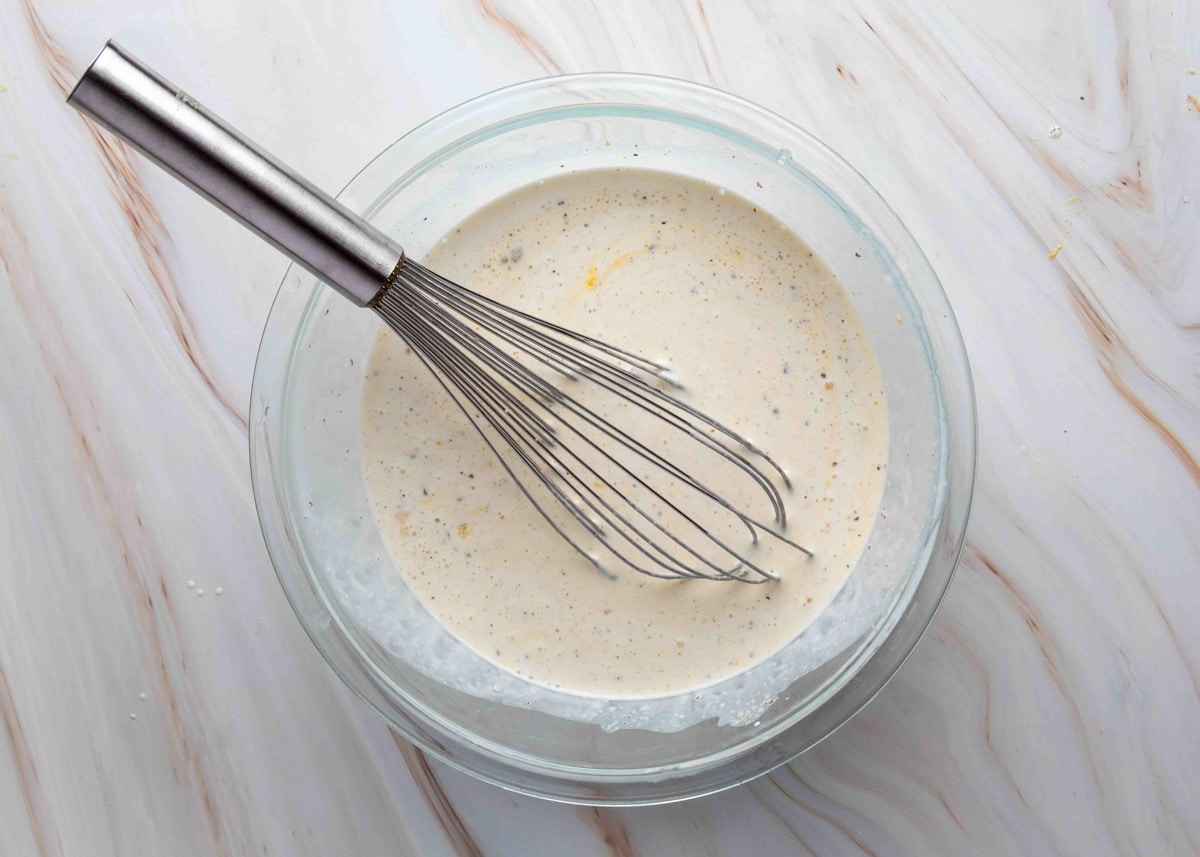 A glass mixing bowl filled with a creamy, speckled egg custard mixture being whisked with a metal balloon whisk, prepared for a sausage croissant breakfast casserole.