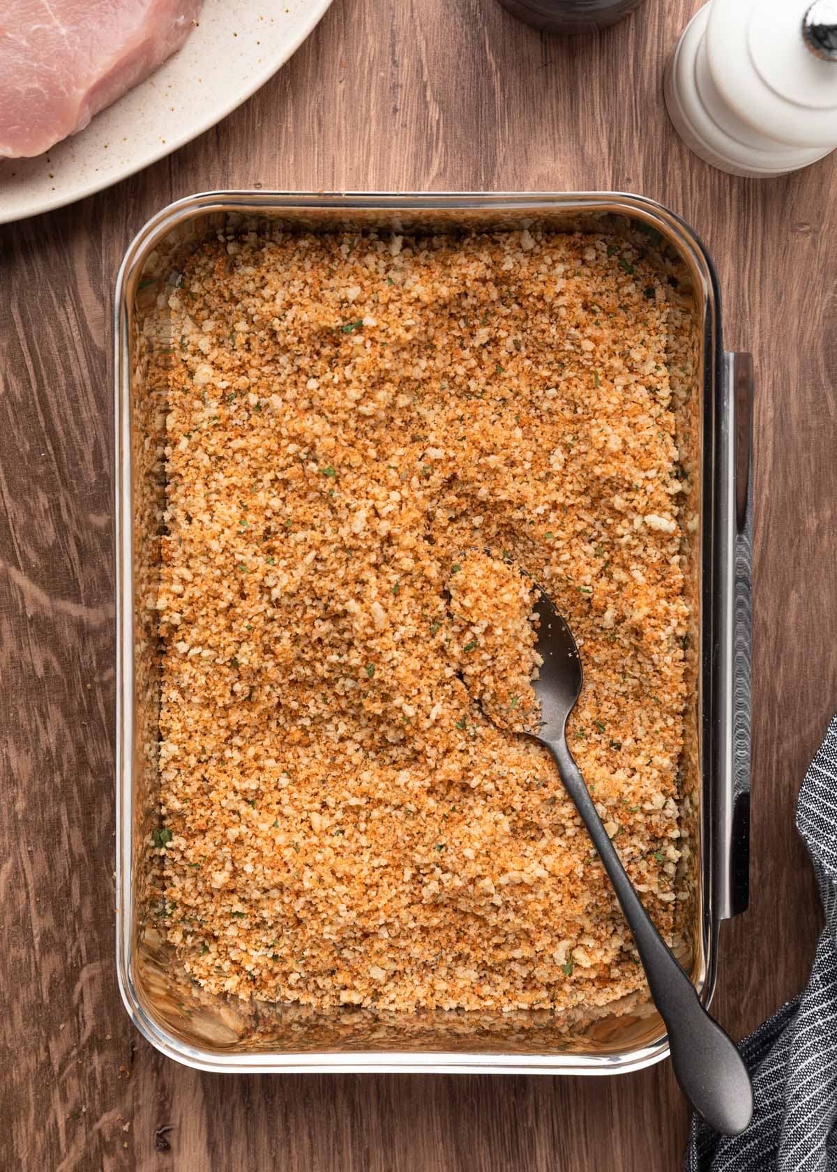 Seasoned panko breadcrumb mixture fully combined with oil in a metal pan, ready for breading pork chops, with a spoon resting on top.