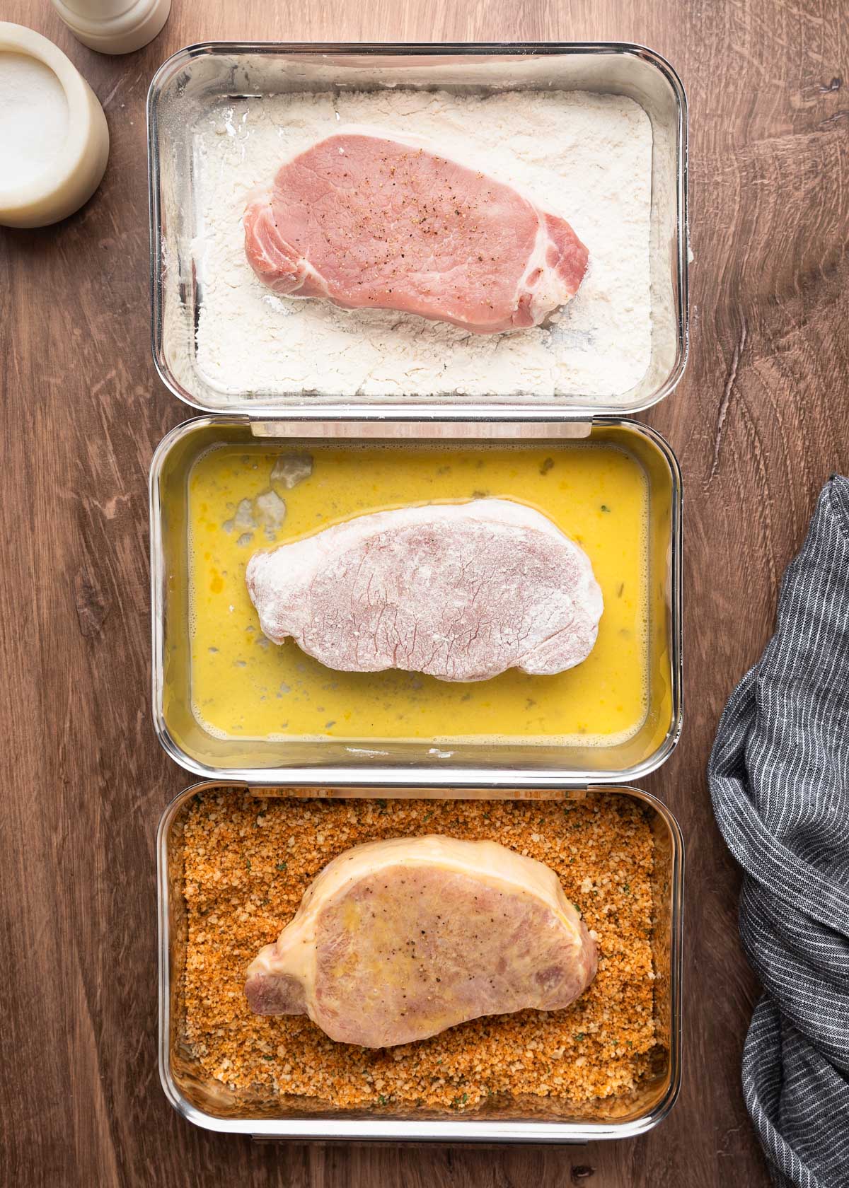 Overhead view of three metal trays in a row showing the breading process for pork chops: the top tray holds a raw pork chop in seasoned flour, the middle tray contains a floured chop in beaten egg, and the bottom tray shows a coated chop resting on seasoned breadcrumbs.