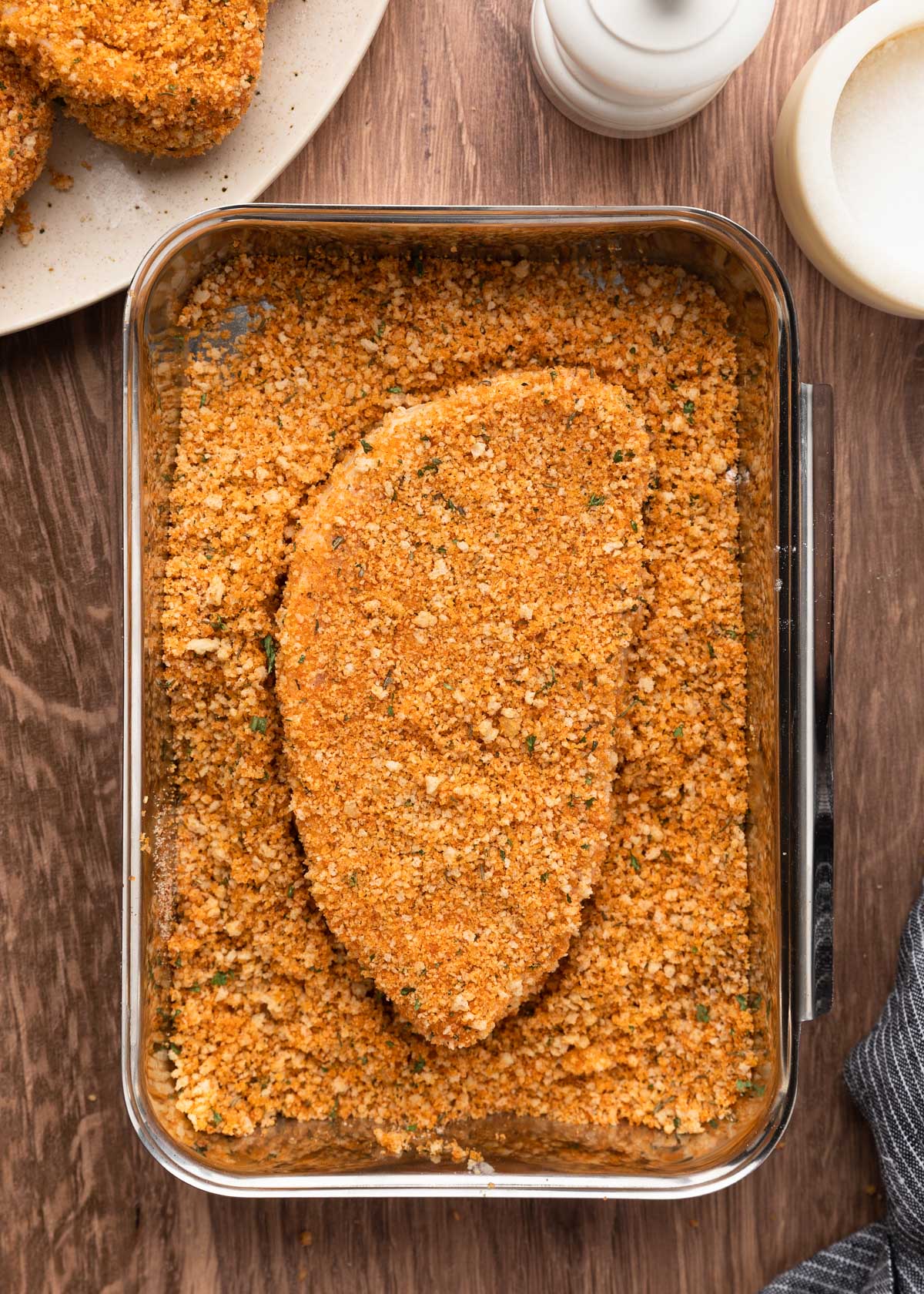 A fully breaded pork chop sits in a tray of golden, seasoned panko breadcrumbs, surrounded by scattered crumbs; part of a plate with more breaded chops is visible in the upper left corner.