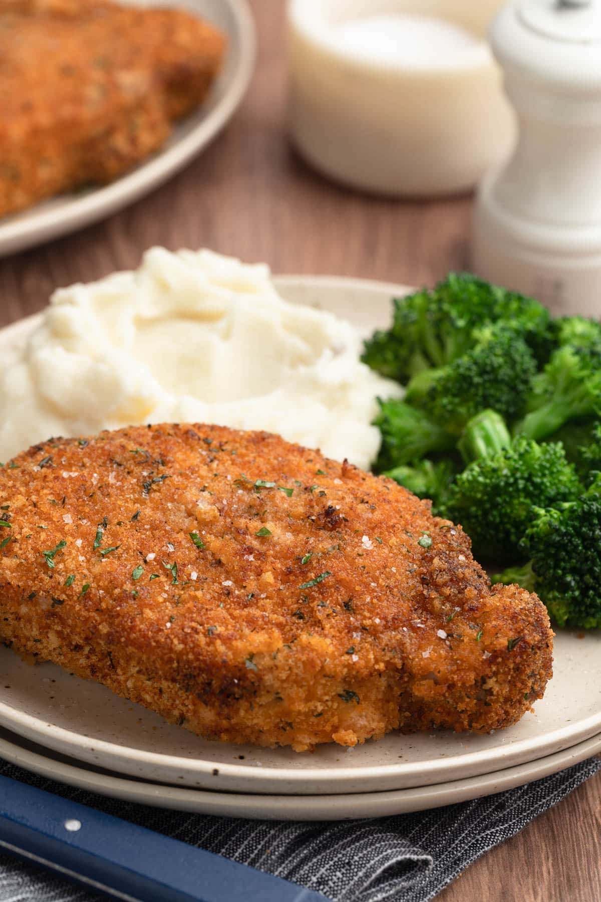 Close-up of a crispy, golden-brown breaded pork chop served on a plate with creamy mashed potatoes and bright green steamed broccoli, with a salt cellar and pepper grinder in the background.