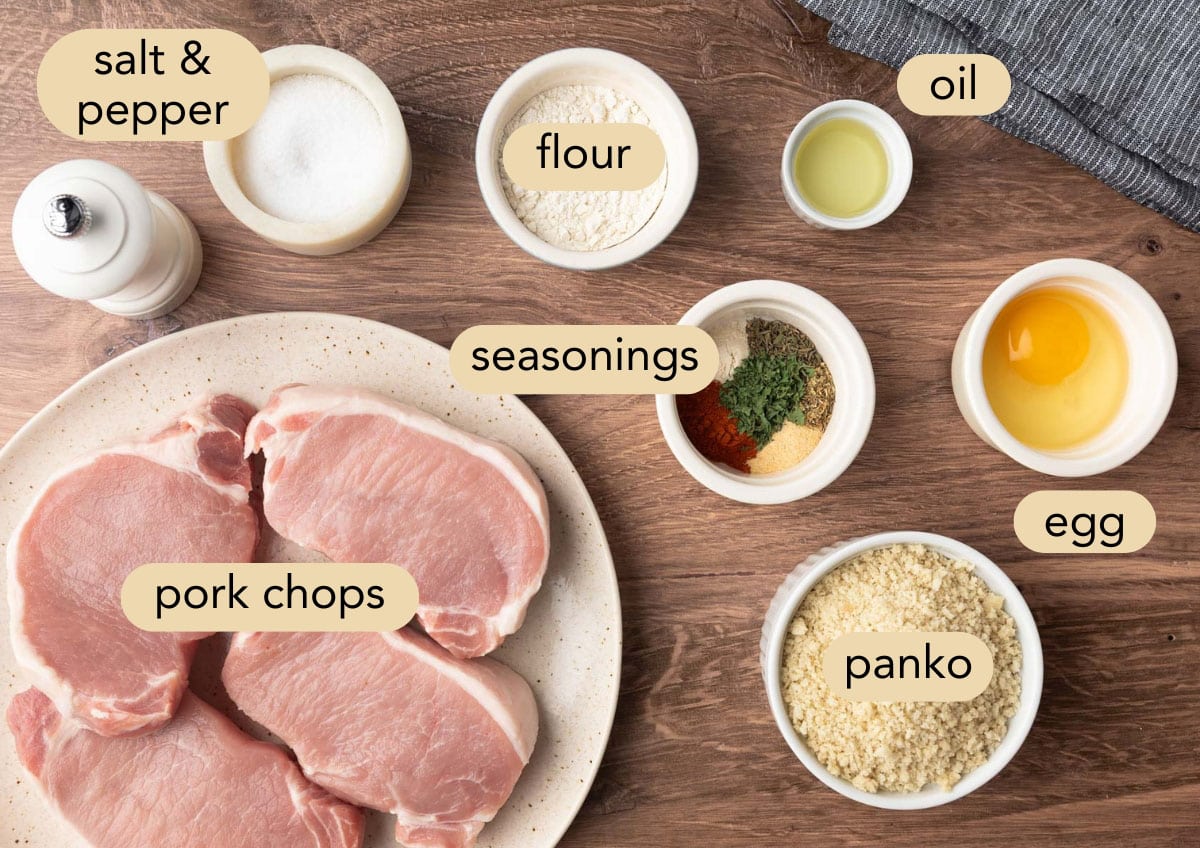 Flat lay of labeled ingredients for air fryer breaded pork chops, including raw pork chops, flour, egg, oil, salt and pepper, panko breadcrumbs, and a small bowl of colorful seasonings on a wood surface.