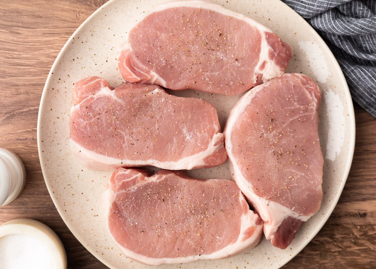 Four raw boneless pork chops on a speckled plate, evenly seasoned with salt and pepper, ready for breading.