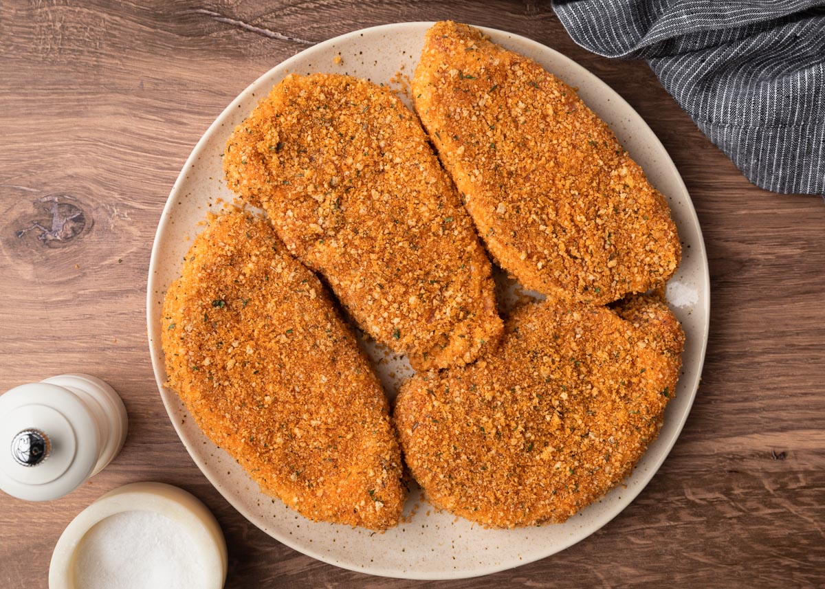 Four pork chops coated in a golden seasoned panko breadcrumb mixture, arranged on a plate before air frying.