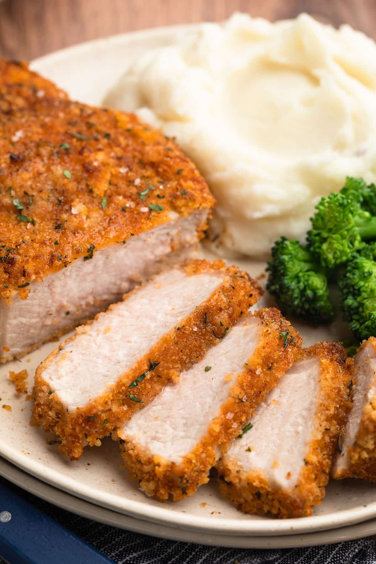 Juicy breaded pork chop sliced to reveal the interior, plated with mashed potatoes and broccoli on a cream-colored plate.