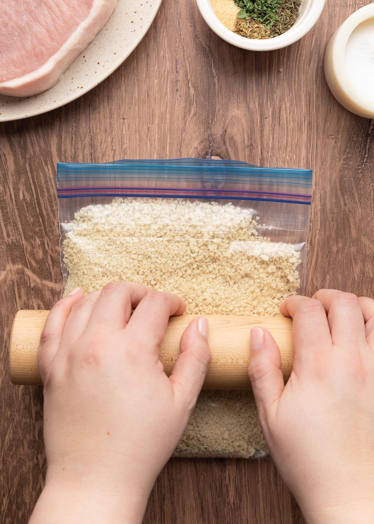Hands using a wooden rolling pin to crush panko breadcrumbs inside a zip-top plastic bag on a wood surface, with ingredients for breaded pork chops visible around the frame.