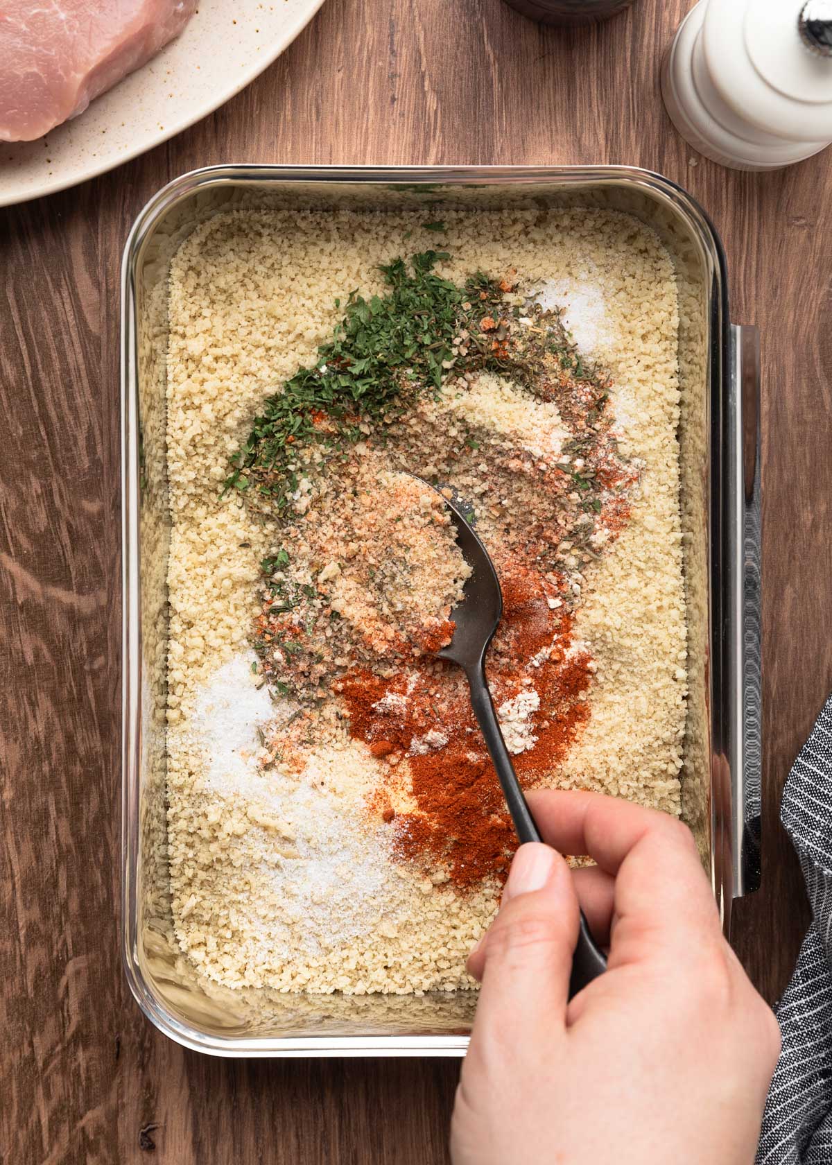 A hand stirring a breadcrumb mixture in a metal tray with a spoon, blending panko with colorful seasonings like paprika, parsley, and garlic powder for air fryer breaded pork chops.